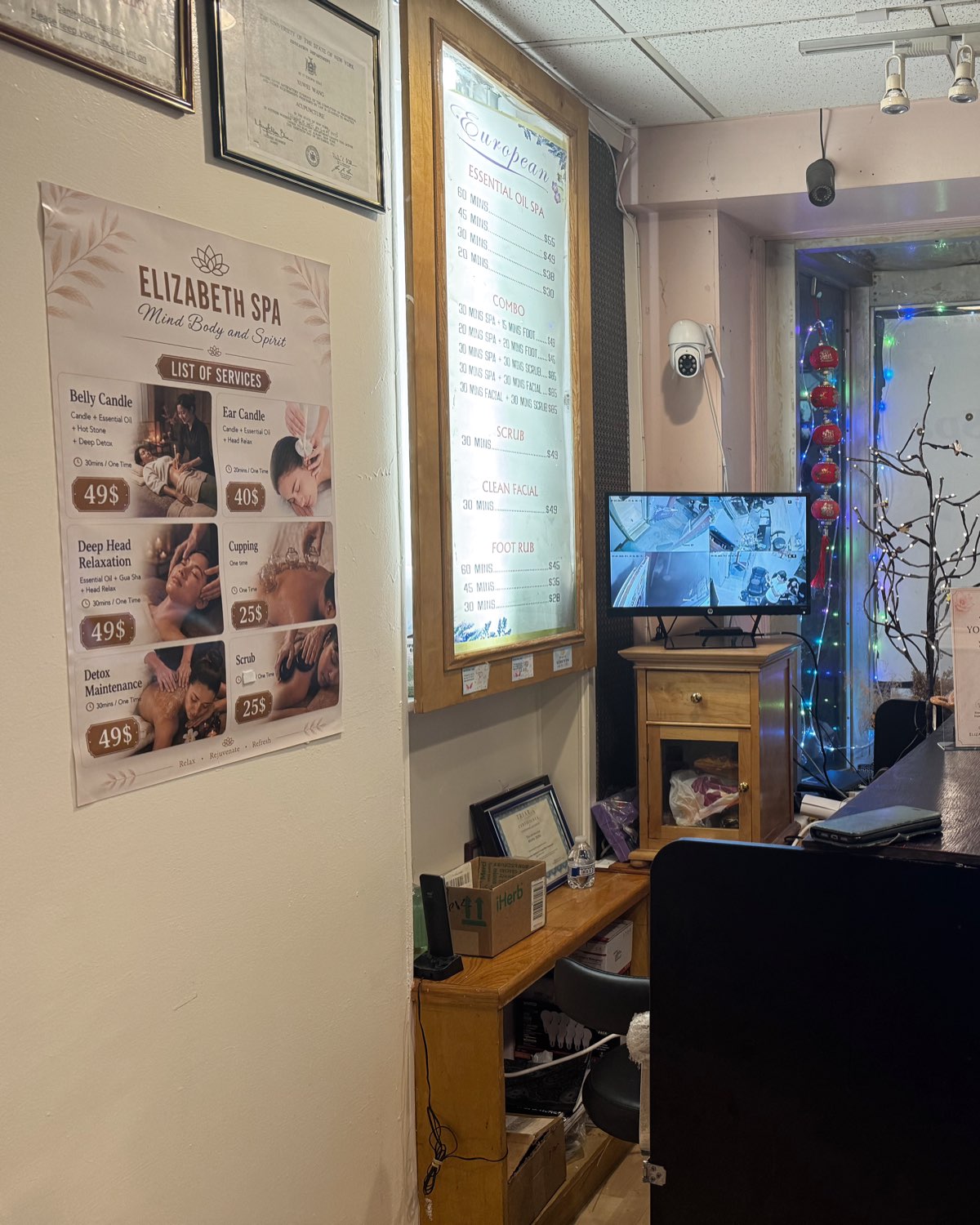 The Elizabeth Spa reception: the printed list of services and the backlit price board on the wall, a wooden desk, framed licences and red lunar-new-year decorations at the window.