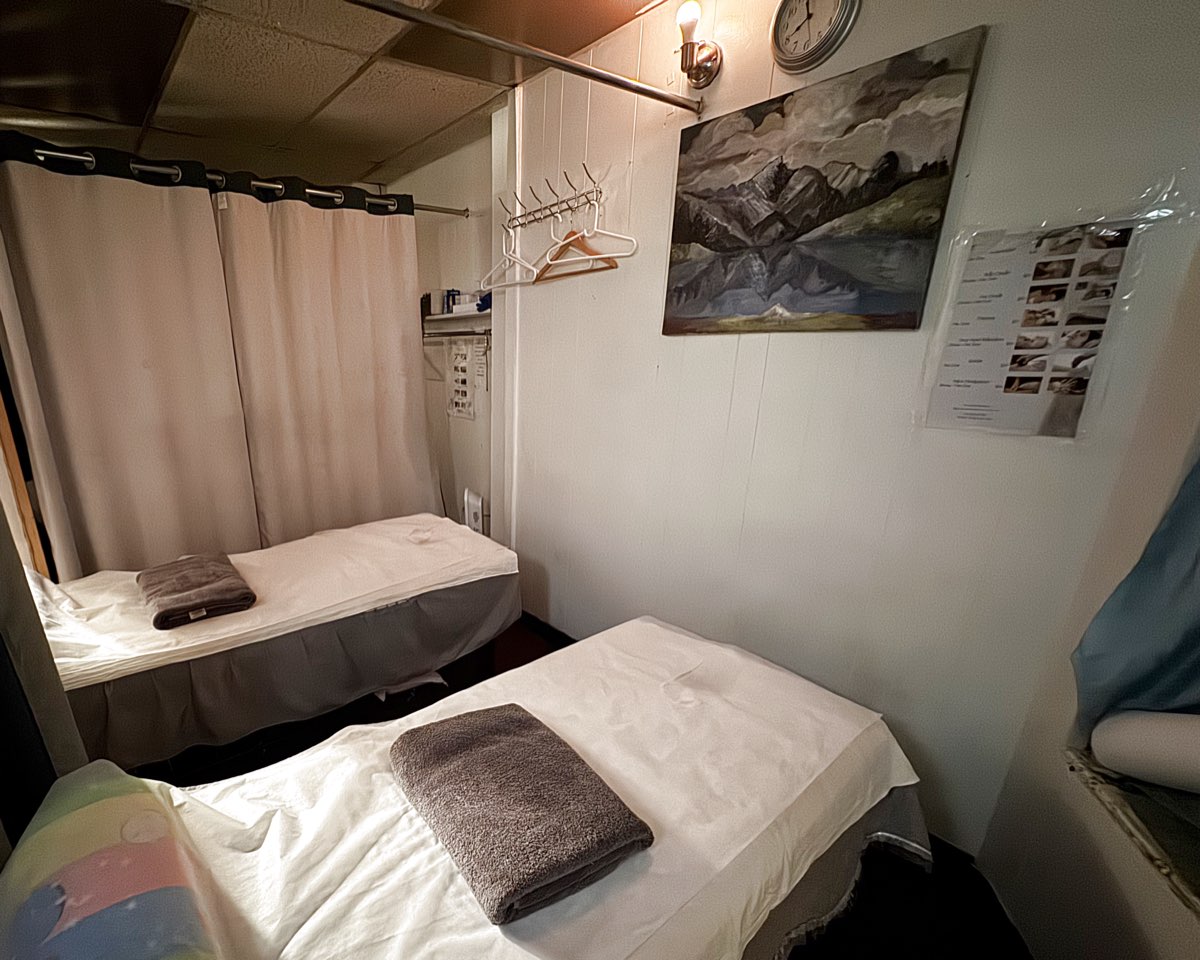 A two-bed treatment room at Elizabeth Spa: two massage tables made up with white linen and folded towels, a curtain divider, coat hangers and a painting of mountains.